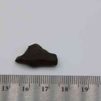 Closeup of basaltic glass flake with ruler: A small, dark basaltic glass flake lies adjacent to a metal ruler, showing its approximate size of around 2 centimeters.