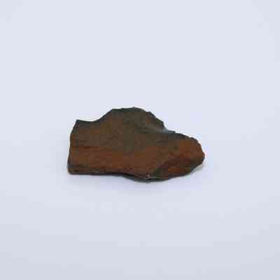 Closeup of basaltic glass flake: The image shows a flake of basaltic glass with a dark, slightly rough texture and an uneven shape, resting on a plain white background.