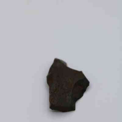 Closeup of basaltic glass flake: A small, dark, jagged rock lies on a plain, light gray surface, its rough texture and irregular shape.