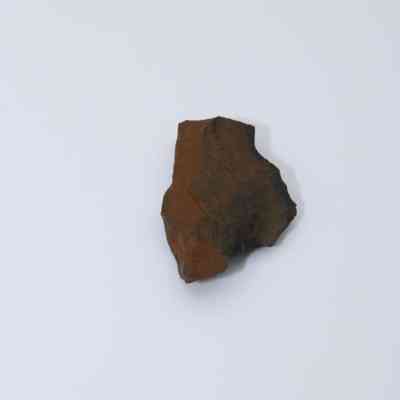 Basaltic glass flake: A dark, irregular basaltic glass flake rests on a smooth, light-colored surface, showcasing distinct textures and rough edges in contrast.