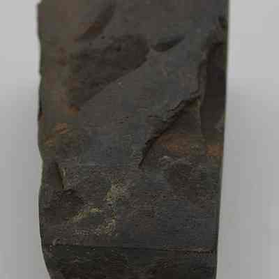 Lengthwise view of basalt adze fragment: The image displays a dark, weathered fragment of an adze, showcasing rough texture and irregular surface, indicative of its age and usage.
