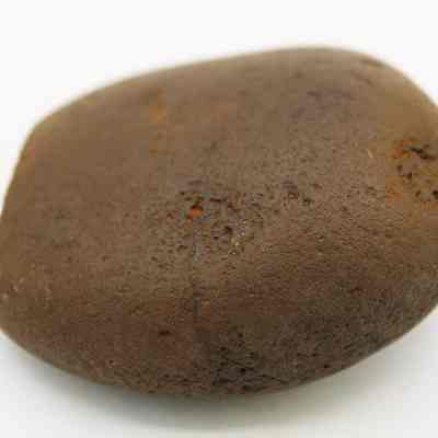 Closeup of basalt hammerstone: The image shows a smooth, brown stone with slight irregularities on its surface, lying on a plain white background. The stone appears oval-shaped.
