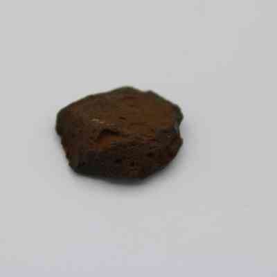 Closeup of glass flake: The image shows a small, dark, irregularly shaped glass flake with a rough texture and earthy color, lying on a plain white background.