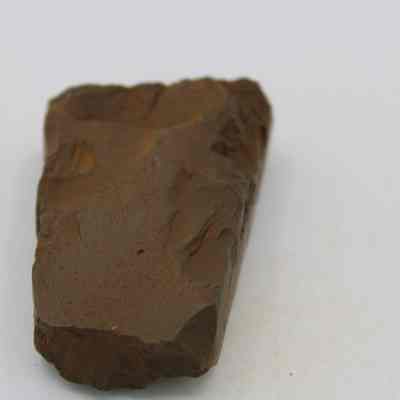 Adze (ko'i) fragment: The image shows a rough, brown stone adze (ko'i) fragment with an uneven surface, resting on a plain white background, displaying various textures and natural indentations.