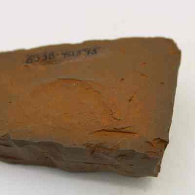 Underside of an adze (ko'i) fragment with item number: The image shows a roughly textured, reddish-brown rock adze (ko'i) fragment with a smooth surface and a sequence of numbers written on it.