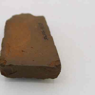 Lengthwise view of the underside of an adze (ko'i) fragment: The image shows a close-up view of a rough, brown rock adze (ko'i) fragment with a handwritten number inscribed on its flat surface.