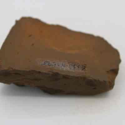 Underside of an adze (ko'i) fragment with item number in black ink: The image shows a rough, brown, rectangular stone adze (ko'i) fragment with a code inscribed on its surface.