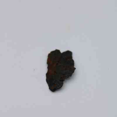 Closeup of basaltic glass flake: A small, dark basaltic glass flake rests on a plain, light gray surface, showcasing its rough texture and irregular shape against a simple backdrop.