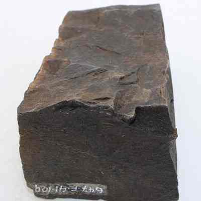 End of basalt adze fragment with original id number visible: The image depicts a rough-hewn fragment of a stone adze, showcasing uneven surfaces and sharp edges, with a number visible near the base.