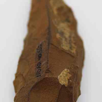 Lengthwise view of basalt awl (kui houhou, kui iwi) with alternate id number visible: An ancient stone tool, likely an awl (kui houhou, kui iwi), is depicted with a sharp, pointed edge and a hand-chiseled, textured surface.