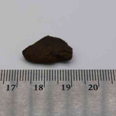 Basaltic glass piece with ruler: A small piece of basaltic glass is placed next to a ruler for scale, showing its size and irregular shape against a plain background.