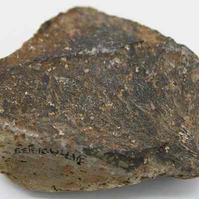 Closeup of basalt scraper with alternate id number visible: The image shows a roughly textured, irregularly shaped rock with a mix of dark and light brown colors, featuring identification number hand-written in black ink (photographed upside down) on its surface.