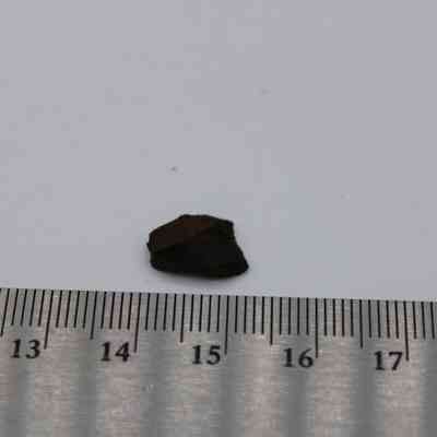 Basaltic glass flake with ruler: A small basaltic glass flake lies next to a ruler on a plain white background, showcasing its size and dark color.