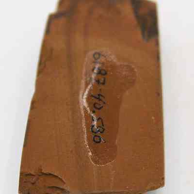 Bottom of small basalt adze (ko'i) with alternate id number visible: The image depicts a close-up view of a worn-out, rectangular stone adze (ko'i) head with a smooth, reddish-brown surface and visible markings.
