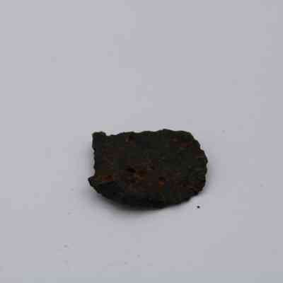 Closeup of volcanic glass flake: The image shows a dark, irregularly shaped volcanic glass flake with a rough texture, resting on a plain white background, highlighting its natural formation.