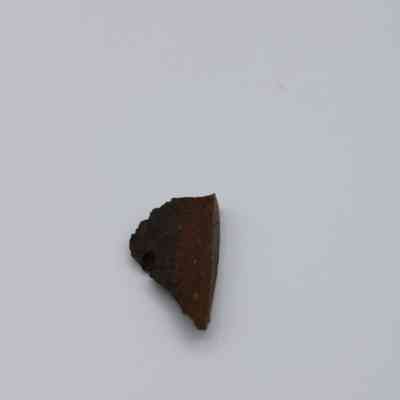 Closeup of basaltic glass flake: A small, dark basaltic glass flake lies on a plain white surface, showcasing its rough texture and irregular edges.