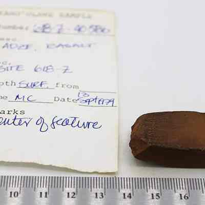 Adze (ko'i) with artifact slip & ruler: The image shows a small, brown, blade-like adze (ko'i) fragment beside a handwritten sample tag, placed on a white surface with a ruler for scale.