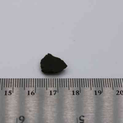Basaltic glass flake with ruler: The image shows a small basaltic glass flake placed beside a ruler, highlighting its size and dark color against a plain background.