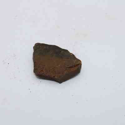 Closeup of basaltic glass flake: A small basaltic glass flake is resting on a light, slightly textured surface, showing its rough, dark appearance with subtle earthy tones.