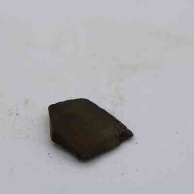 Closeup of basaltic glass flake: A flat, dark basaltic glass flake rests on a light surface with scattered small particles nearby, appearing matte and irregularly shaped.