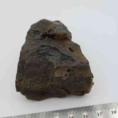 Closeup of basaltic glass core with ruler: A rough, dark basaltic glass core is positioned in front of a ruler, highlighting its textured surface and varied edges.