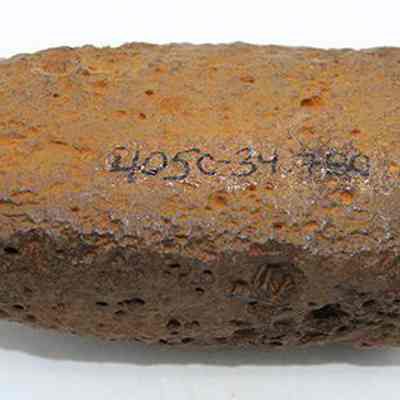 Profile of adze preform with alternate id number visible: The image displays a piece of stone, exhibiting a rough, pitted texture and an irregular shape. A sequence of numbers and letters are written in black in on one side.