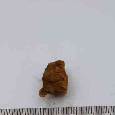 Extreme closeup of a piece of calcite with a ruler: The image shows a small, brown calcite rock fragment resting on a white surface, with a nearby ruler for scale, indicating its compact size.
