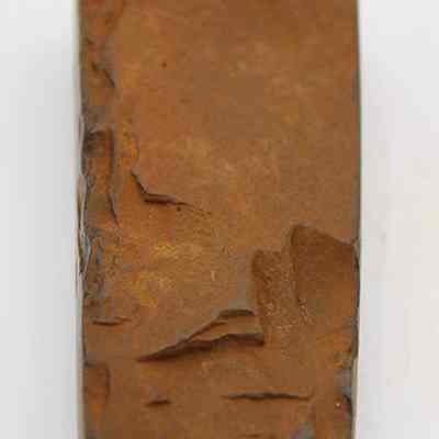 Lengthwise view of unpolished side of basalt adze (ko'i) fragment: The image shows a fragment of an ancient adze (ko'i), featuring a rough, rust-colored surface with visible chipping and wear.
