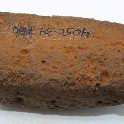 Adze preform with alternate id number is visible: The image shows a pitted, rock with a rough texture and some numbers and letters handwritten in black ink (upside down), placed against a plain white background.