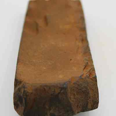 Lengthwise view of polished basalt adze (ko'i) fragment: The image shows a brown, rectangular adze (ko'i) fragment with rough, uneven edges, intricately carved from stone for woodworking purposes.