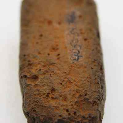Lengthwise view of an adze preform with alternate id number visible: The image features a weathered, rectangular, brown-colored rock with a rough texture and visible pockmarks, possibly inscribed with faint markings or symbols
