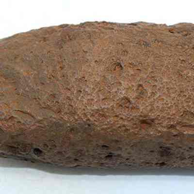Adze preform: The image shows a brown, rough-textured, irregularly-shaped rock resting on a plain white background. The rock has a pitted surface.