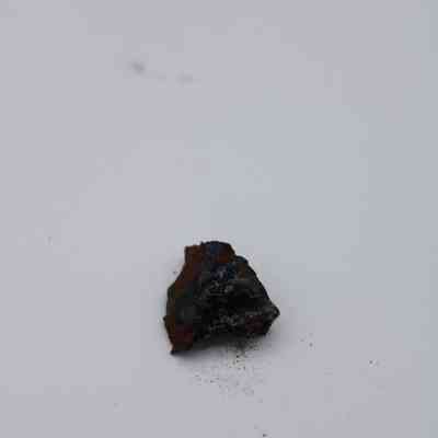 Closeup of basaltic glass core: A rough, dark basaltic glass core fragment rests on a plain surface, surrounded by scattered specks of reddish-brown and black debris.