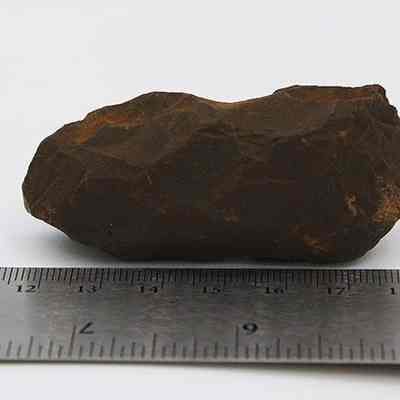 Profile of adze preform: The image depicts the profile of a stone adze preform, showing its rough, chipped surface, placed next to a ruler for size reference on a white background.