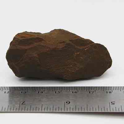 Reverse side of adze preform: The image depicts a rough, brown adze preform stone lying next to a ruler, measuring approximately 6 centimeters in length on a neutral background.