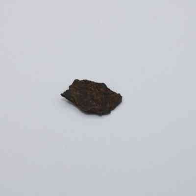 Closeup of tiny basaltic glass flake: A small, dark basaltic glass flake rests on a plain, light surface, showcasing its textured and jagged edges.