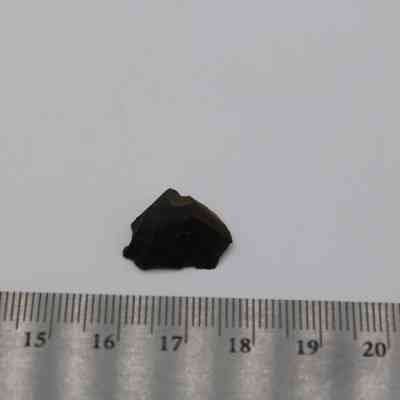 Closeup of glass flake with ruler: A small, dark glass flake is placed on a white surface next to a metal ruler, indicating its approximate size.