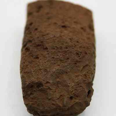 Lengthwise view of an adze preform: The image features a rough, brown stone artifact with an elongated, slightly rounded shape, textured surface, displaying various indentations and irregularities.