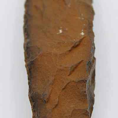 Lengthwise view of quadrangular adze: The image shows a weathered, brown stone tool with sharp edges, likely used historically for cutting purposes, resting on a plain white background.