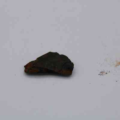 Closeup of basaltic glass flake: A small, dark basaltic glass flake lies on a light surface, surrounded by scattered fine particles.