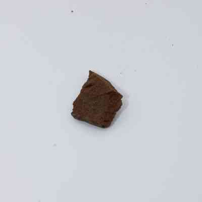 Closeup of basaltic glass flake: The image shows a small, rough-textured basaltic glass flake with a brownish color, resting on a smooth, light background.