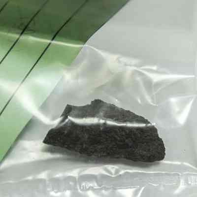 Basalt flake in a plastic bag: A small, dark rock fragment is sealed inside a transparent plastic bag, partially visible under a sheet of green paper with lines.