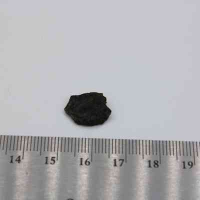 Small basaltic glass flake with ruler: The image shows a small basaltic glass flake next to a metal ruler, measuring approximately 1.5 cm in length, placed on a white surface.
