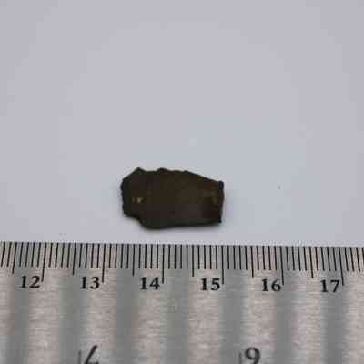 Closeup of small basaltic glass flake with ruler: A small, dark basaltic glass flake is placed on a white surface next to a ruler for scale reference.