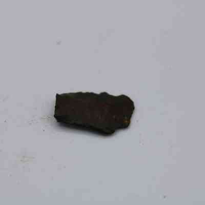 Closeup of basaltic glass flake: The image displays a solitary, dark basaltic glass flake resting on a light surface, with fine, scattered particles surrounding it.