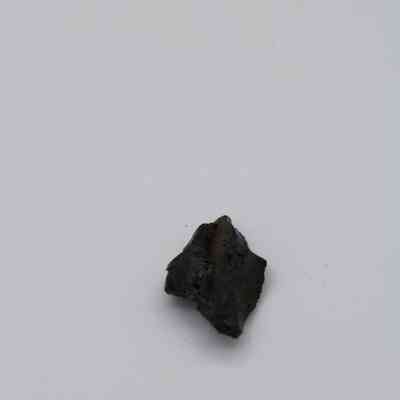 Closeup fo basaltic glass cortex flake: A small, dark basaltic glass cortex flake rests on a pale, featureless background, showcasing its rough texture and irregular shape.