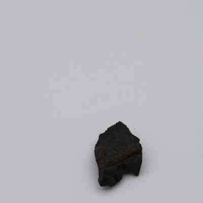 Closeup of basaltic glass cortex flake: The image depicts a small basaltic glass cortex flake resting on a plain white background, showcasing its dark, rugged texture.
