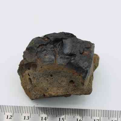 Closeup of basaltic glass core with ruler: The image shows a piece of basaltic glass core with a rough, dark surface and some textured areas, placed next to a ruler.
