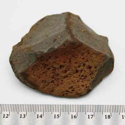 Close up of core/scraper and ruler: The image shows a roughly textured, reddish-brown rock with dark edges, placed next to a ruler for scale measurement.
