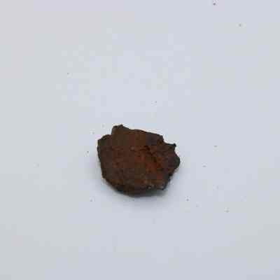 Tiny piece of basaltic glass: The image shows a small, irregularly shaped piece of basaltic glass with a dark, earthy appearance on a white surface.